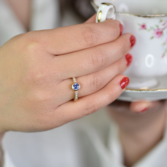 frau hält teetasse und zeigt Gelbgold Verlobungsring mit Pavé-Fassung – goldring 750 mit diamanten und blauem Saphir, elegant getragen.