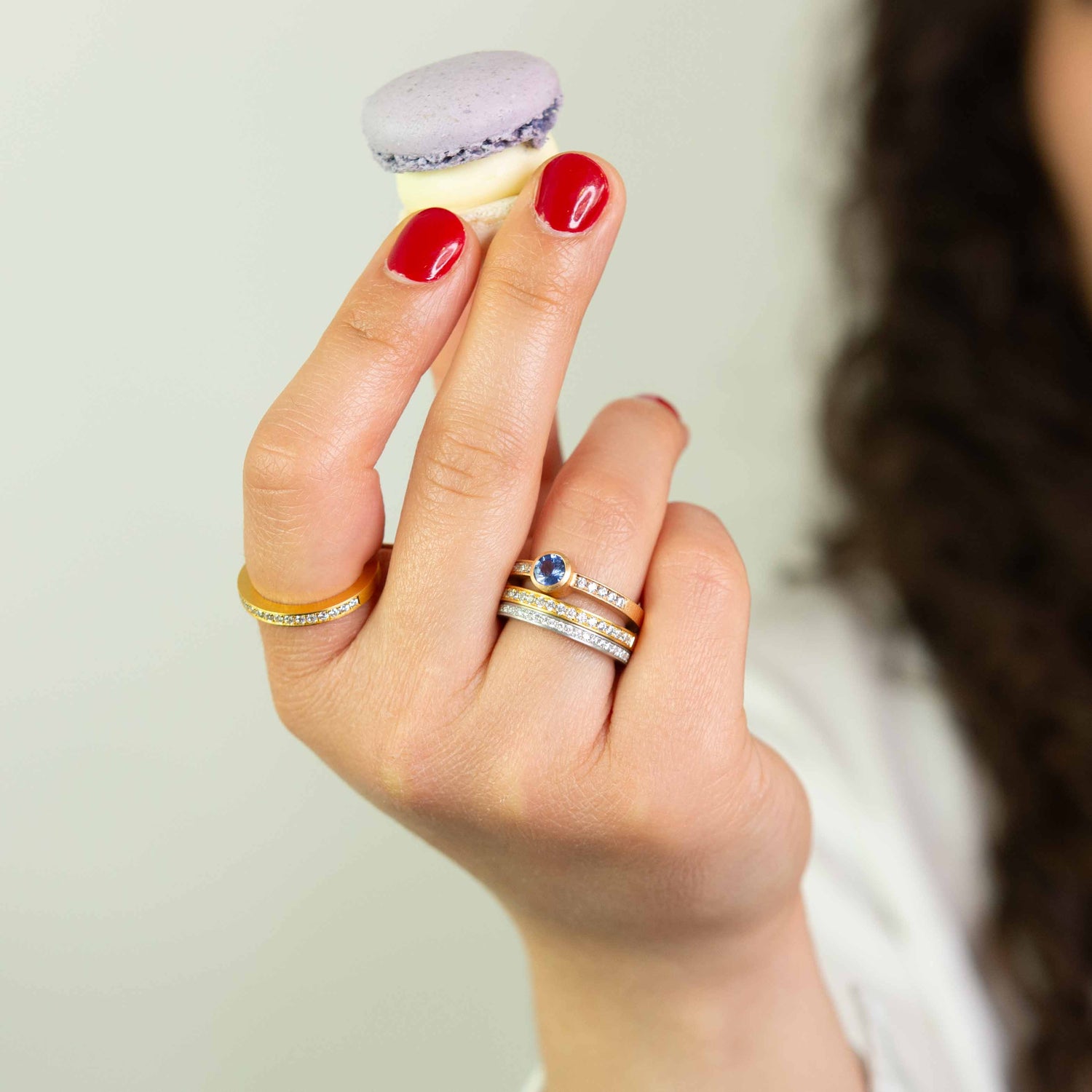 Frau hält Macaron und trägt gelbgoldenen Pavé-Ring mit Saphir und Diamanten sowie weitere Ringe mit diamanten rundherum